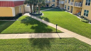 A courtyard with a well-maintained lawn, a central walkway, and a building with multiple balconies and windows. - Ocean View  Second Floor 2/2 (New Smyrna Beach)