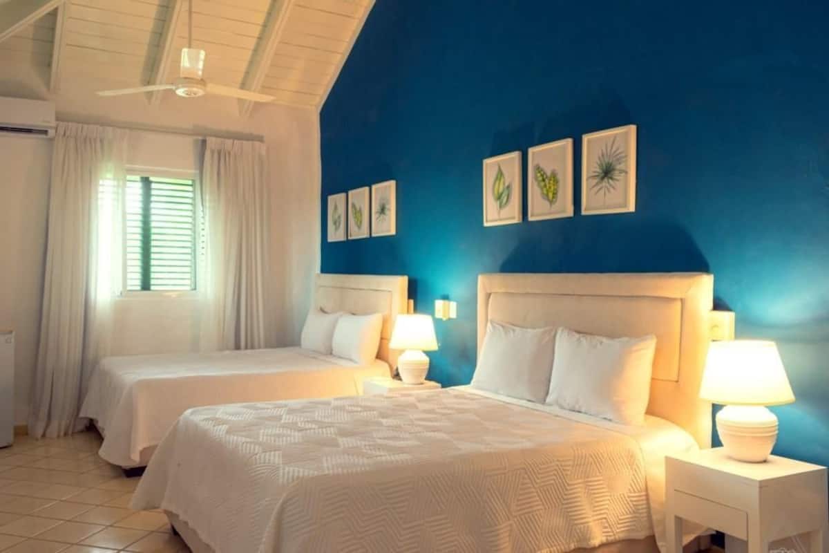 Habitación de hotel con dos camas, ventilador de techo, ventana con cortinas y cuadros colgados en la pared.
