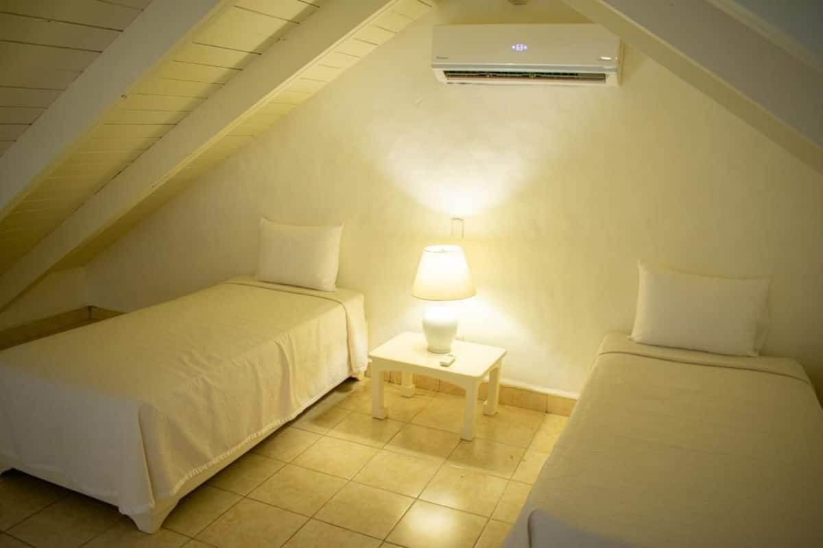 Una habitación pequeña y acogedora con dos camas individuales, una mesita de noche con lámpara y un equipo de aire acondicionado en el techo inclinado.