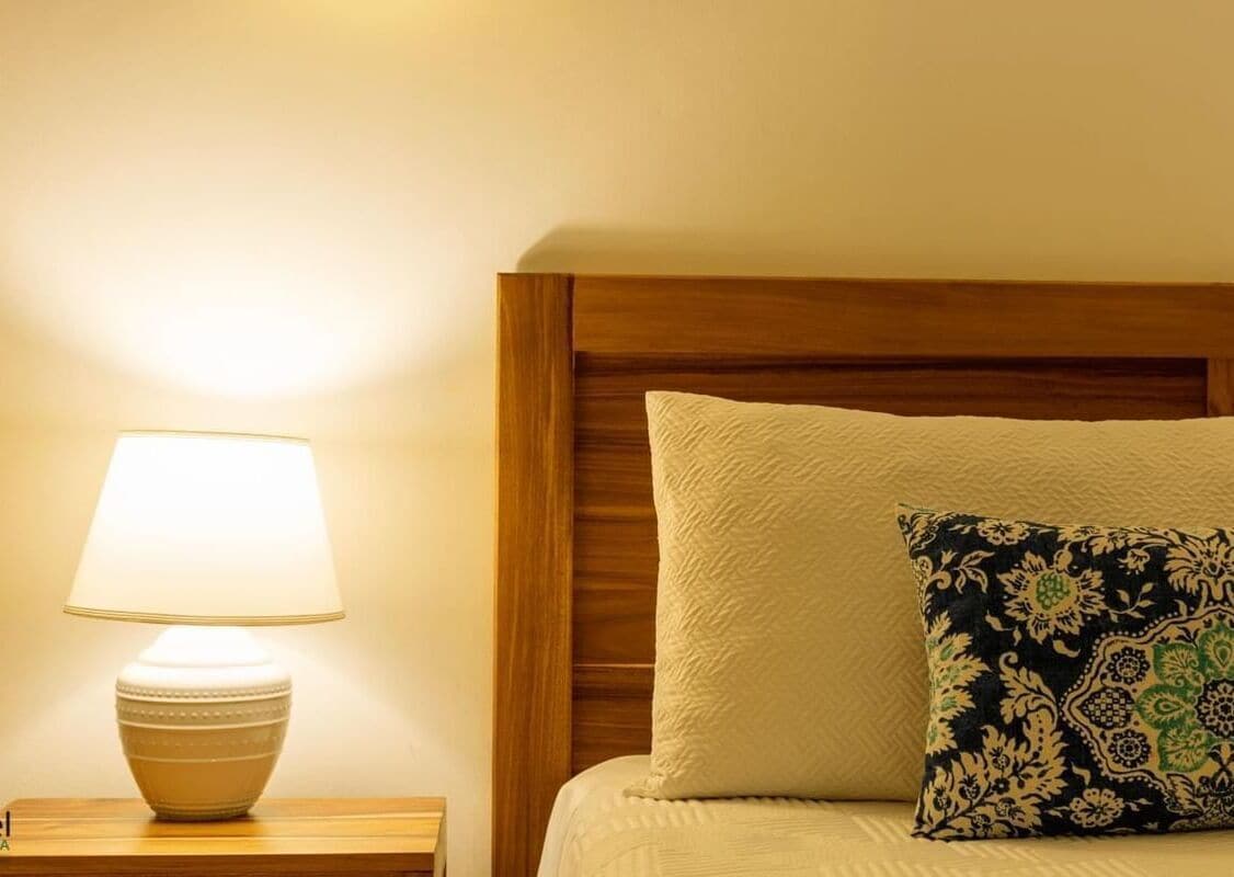 Una cama bien hecha con un almohadón y una lámpara de noche sobre una mesita de madera.
