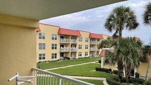 A balcony with a view of a multi-story building with balconies, a lawn, and palm trees.