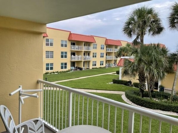A balcony with a view of a multi-story building with balconies, a lawn, and palm trees.