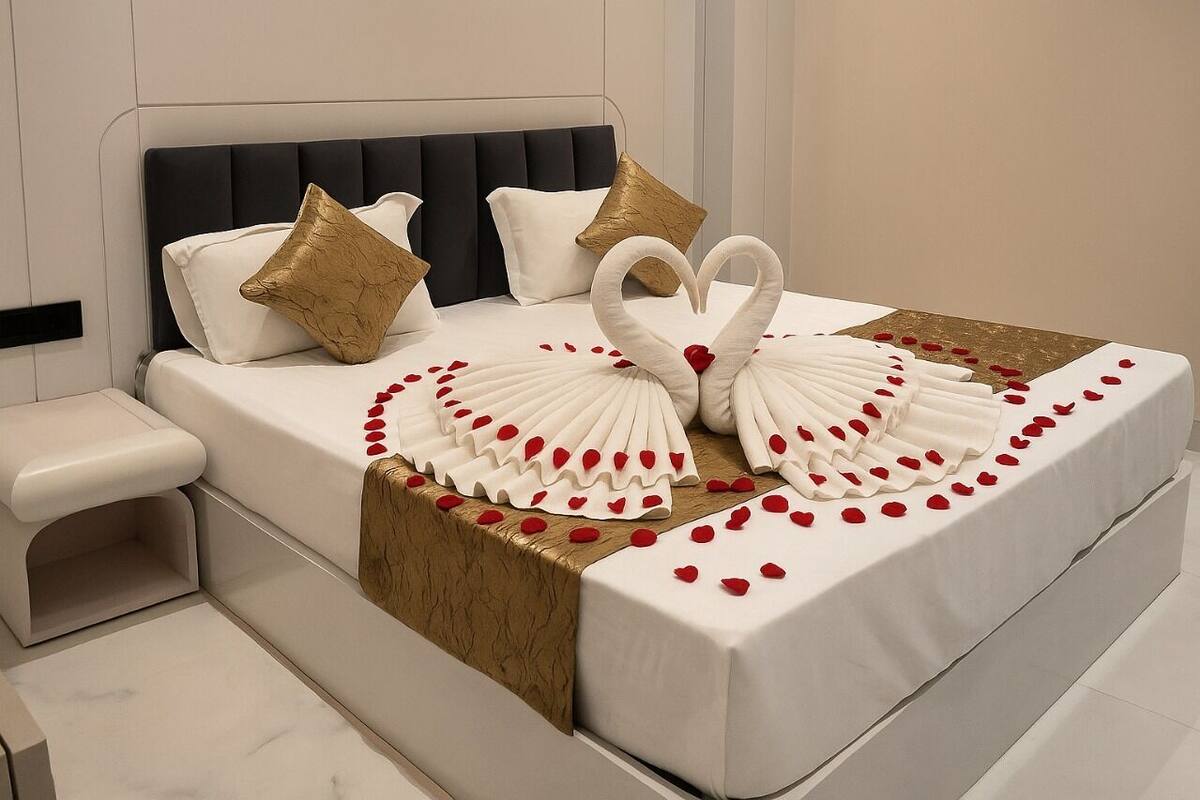 A neatly made bed with a heart-shaped swan decoration and rose petals, a bedside table, and a headboard.