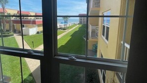 A view from a window overlooking a well-maintained lawn, a building with balconies, and a distant beach. - Ocean View Second Floor 2/2 (New Smyrna Beach)