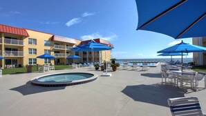 Un área de piscina con sillones de descanso y sombrillas, una piscina y un edificio con balcones.
