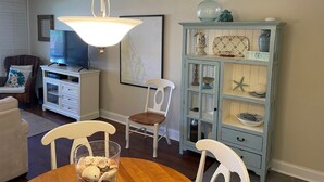 A dining area with a wooden table, white chairs, a glass vase with seashells, a light blue cabinet with decorative items, and a television on a white cabinet. - Ocean View Second Floor 2/2 (New Smyrna Beach)
