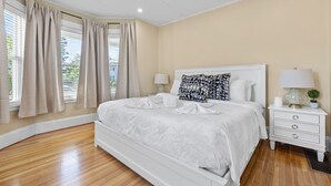 Un dormitorio con una cama grande, mesitas de noche blancas, paredes color beige y una ventana con cortinas.