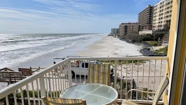 A balcony with a glass table and striped chairs overlooking a beach with waves and buildings in the background. - Ocean View Third Floor 2/2 (New Smyrna Beach)