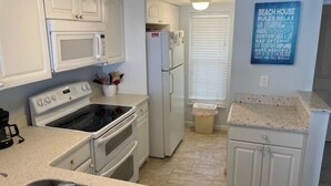A kitchen with white cabinets, a microwave, stove, and a refrigerator. There is a trash can and a window with blinds. A sign on the wall reads "Beach House Rules Relax". - Ocean View Third Floor 2/2 (New Smyrna Beach)