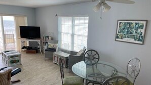 A living room with a glass dining table, wicker furniture, a ceiling fan, and a large window with blinds. - Ocean View Third Floor 2/2 (New Smyrna Beach)