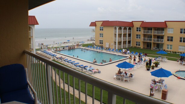 A hotel pool area with a view of the beach, lounge chairs, umbrellas, and a building with a red roof.