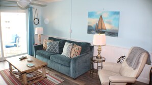 A living room with a teal sofa, a wooden coffee table, a beige armchair, and a painting of a sailboat on the wall.