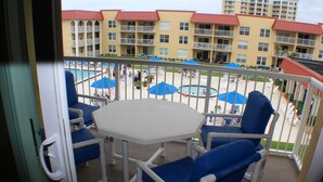 A balcony with a white table and blue chairs, overlooking a pool area with umbrellas and a building with balconies in the background.