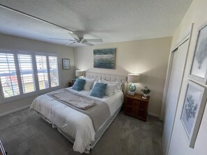 A bedroom with a bed, a ceiling fan, a nightstand with a lamp, and a window with blinds.