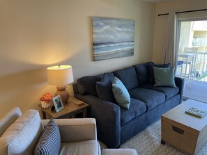 A living room with a sofa, a coffee table, a lamp, a small table with decorative items, and a painting of a beach scene.
