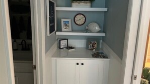 Un petit espace clos comprenant une armoire blanche, une horloge, un tableau encadré et une petite étagère avec une plante.