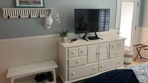 Une chambre avec une commode blanche, un téléviseur posé dessus, une petite étagère blanche ornée d’un cadre photo et un banc sous lequel se trouvent des chaussures.