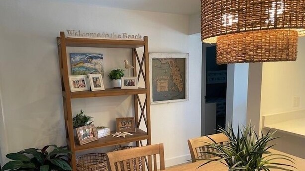Un coin repas avec une table en bois, des chaises et un lustre en osier. Une étagère fixée au mur expose des photos encadrées et une carte.