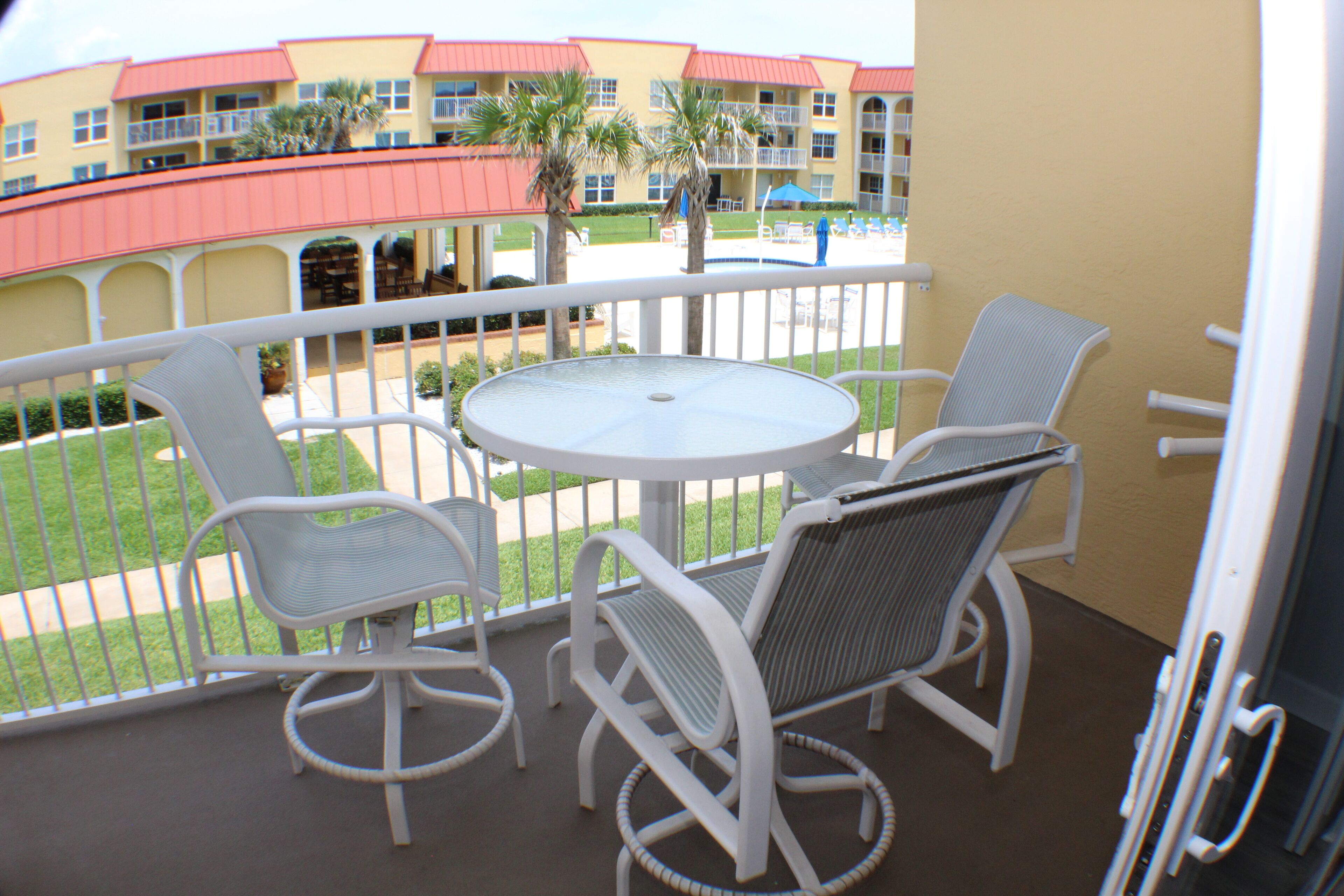 A balcony with a round table and four chairs, overlooking a courtyard with a pool and a building with red roofs.