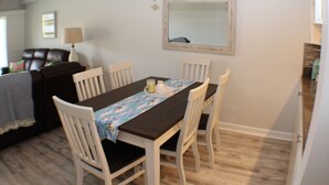 Un comedor con una mesa dispuesta para cuatro personas, rodeada de sillas blancas. Un mantel de madera con estampado floral se coloca sobre la mesa. Hay un espejo en la pared sobre la mesa.