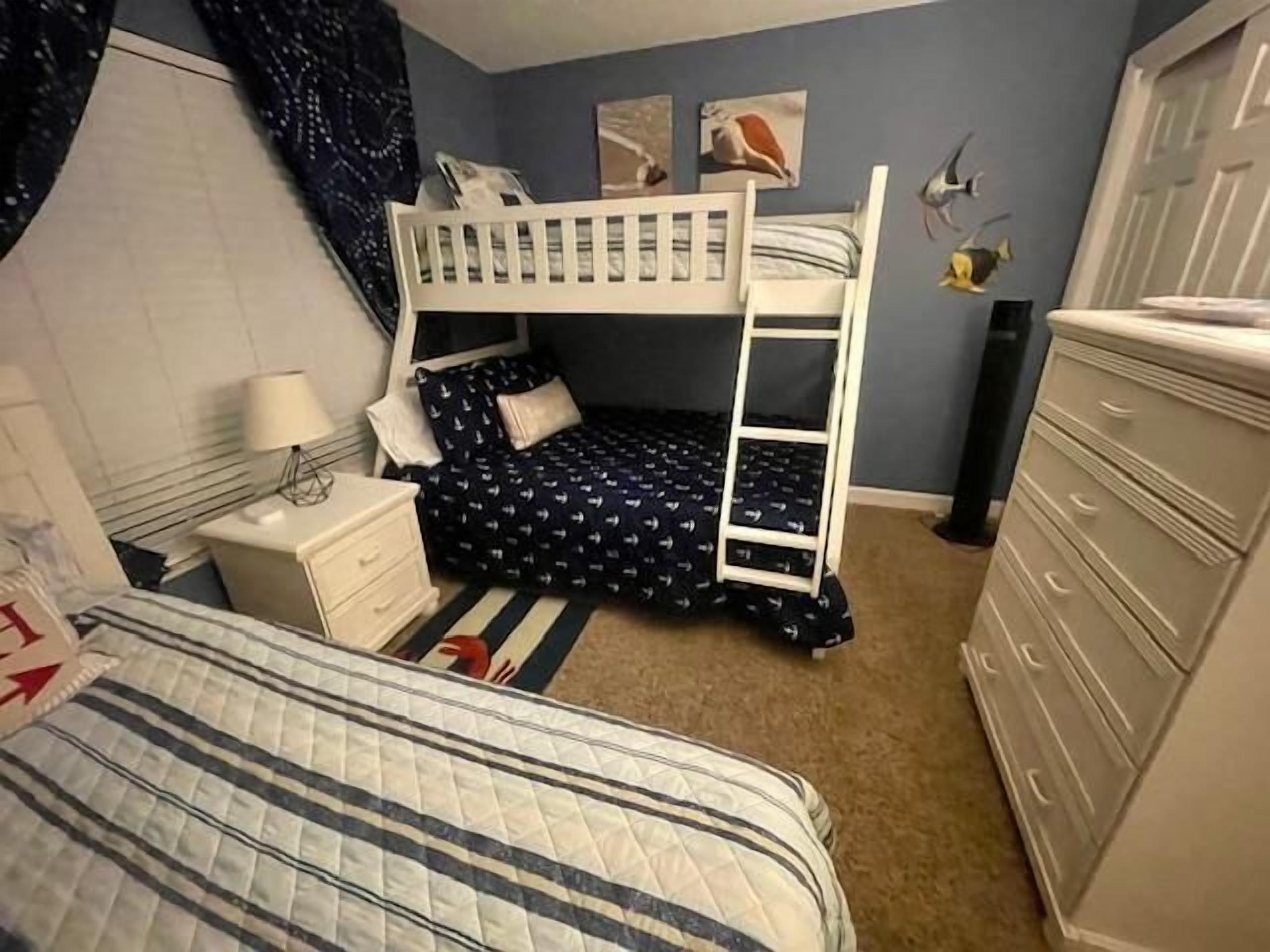 Un lit superposé avec une échelle, une table de chevet avec une lampe, une commode et une couvre-lit à motifs.