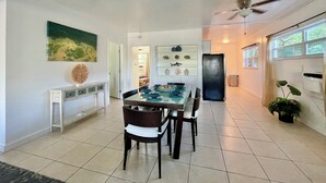 Un vaste espace dédié à la salle à manger, avec une table et des chaises en verre, un ventilateur de plafond et, au fond, une vue sur la cuisine.