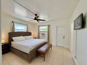 Ein Hotelzimmer mit Bett, Deckenventilator, Bank, Nachttisch, an der Wand montiertem Fernseher und einer Tür.
