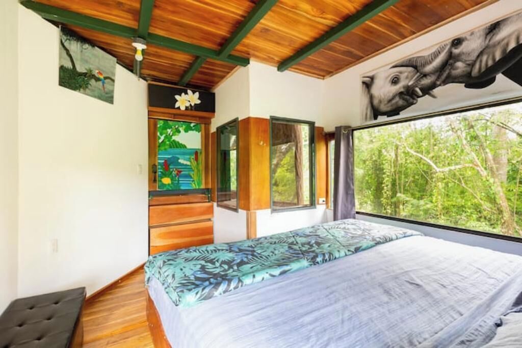 A bedroom with a bed, a window with a view of trees, a wooden ceiling, and a painting on the wall.