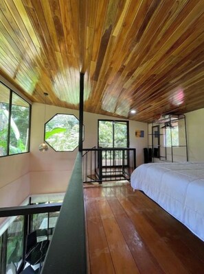 A room with a wooden ceiling, a bed, a balcony, and large windows.