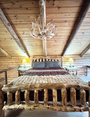 A wooden cabin with a bed featuring antler headboard, two bedside lamps, and a rustic wooden ceiling.