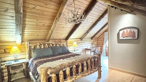 A rustic wooden room with a bed, a wooden headboard, a chandelier, a wall-mounted picture, and a wooden floor. - The Cooperstown Cabin, 8min to All Star Village, 15min to Dreams Park, Sleeps 10 (Oneonta)