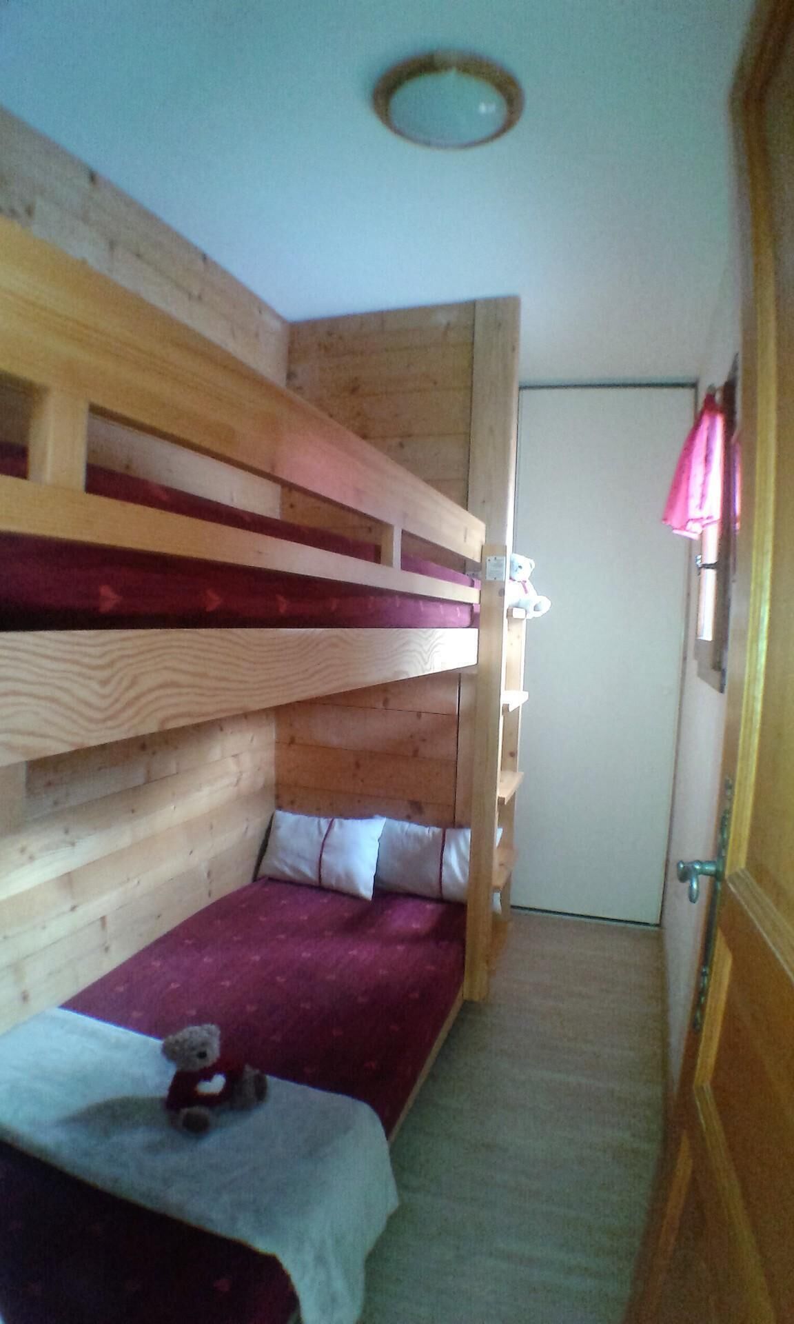 Ein kleiner, schmaler Raum mit einem Etagenbett und einem Einzelbett. Auf dem Einzelbett liegt ein Teddybär.