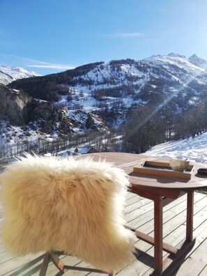 Una terraza de madera con una silla rústica y una mesa, con vistas a un paisaje nevado de montañas.