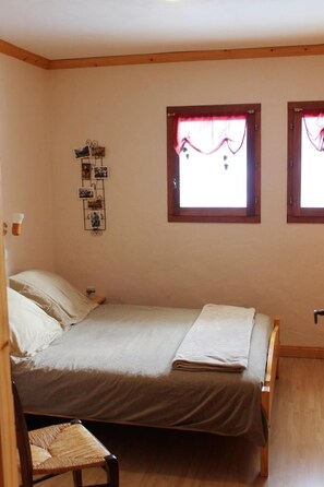Un dormitorio pequeño y acogedor con una cama individual, una mesita de noche, una silla y dos ventanas con cortinas.