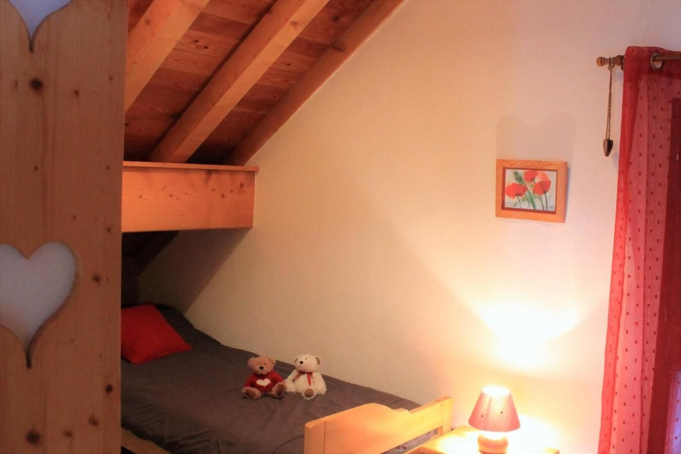 A cozy attic bedroom with a sloped ceiling, a bed with a red pillow, two teddy bears, a small framed picture, and a lamp.