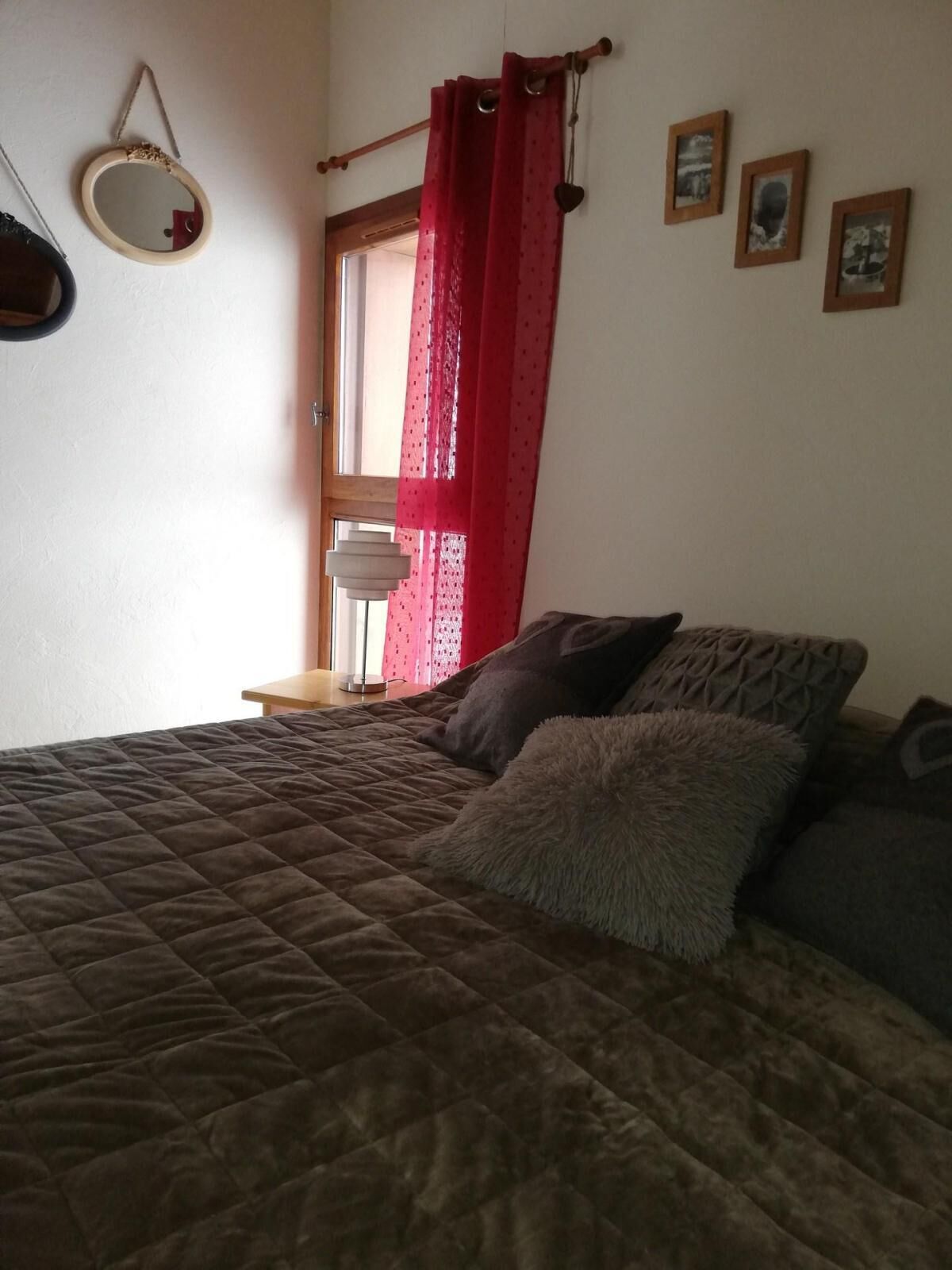 A bedroom with a bed, pillows, a mirror, and a window with red curtains.