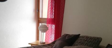 A bedroom with a bed, pillows, a mirror, and a window with red curtains.