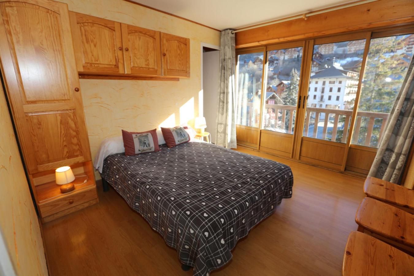 A bedroom with a large bed, wooden cabinets, a nightstand with a lamp, and a window with a view of buildings.