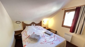 A small, cozy bedroom with a single bed, a nightstand, a wall-mounted lamp, and a window with curtains.