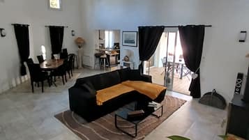 Een ruime woonkamer met een zwarte bank, een eettafel met stoelen en een keuken op de achtergrond.