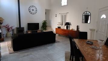 Een ruime woonkamer met een hoge nok, een open haard, een zwarte bank, een houten eettafel en een groot raam dat voor natuurlijk licht zorgt.