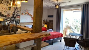 A modern room with a wooden bench, a bed with a red blanket, a dining area, and a balcony with wicker chairs. - A balcony overlooking the magnificent ochre cliffs of Roussillon (Roussillon)