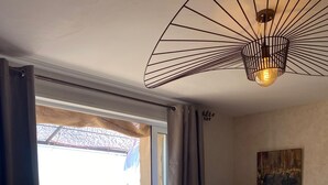 A room with a bed, a small table, a chair, a hanging light fixture, and a view of a balcony with outdoor furniture. - A balcony overlooking the magnificent ochre cliffs of Roussillon (Roussillon)
