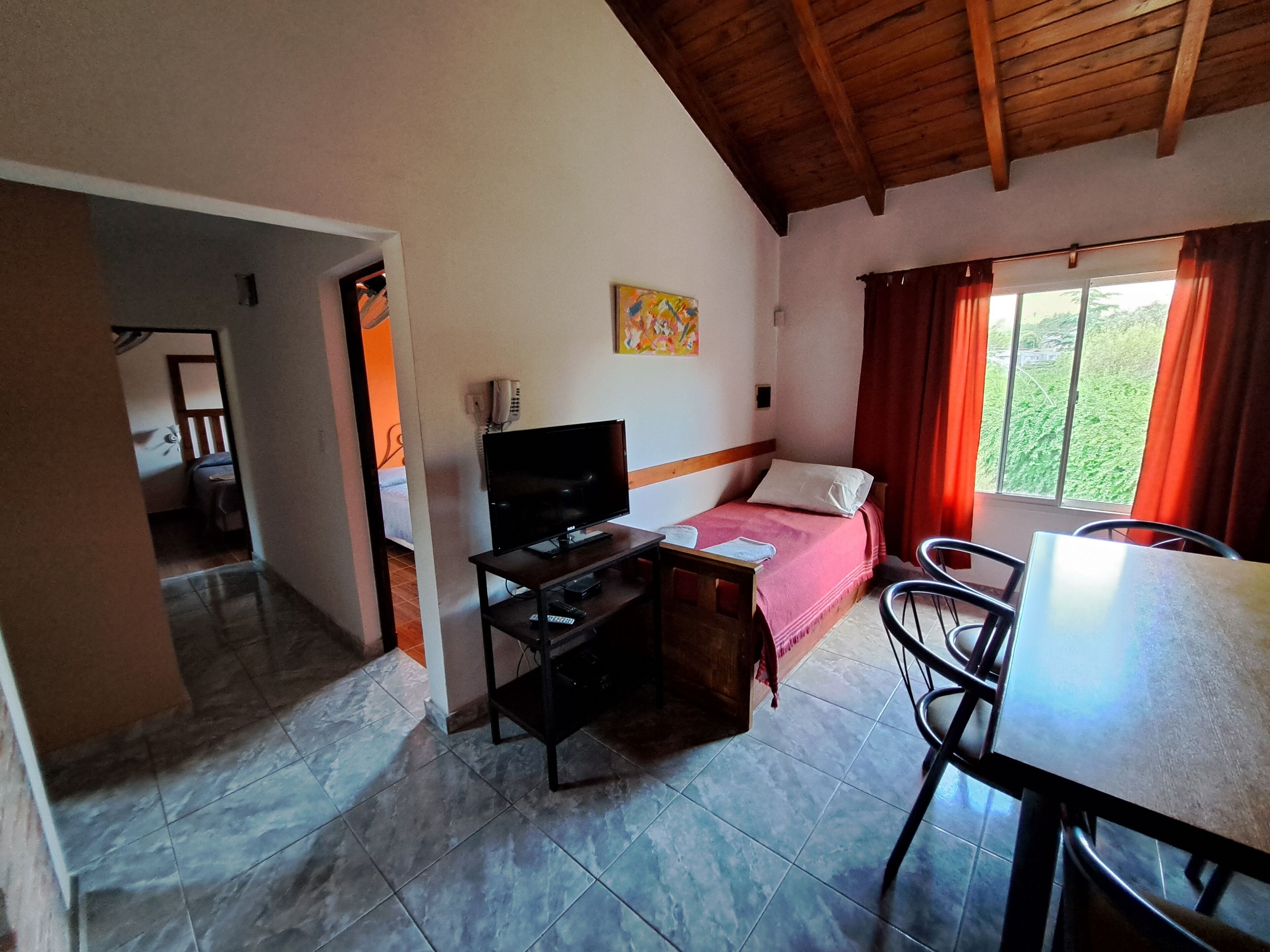 A room with a bed, a television, a desk, and a chair. There is a window with curtains and a view of greenery outside.