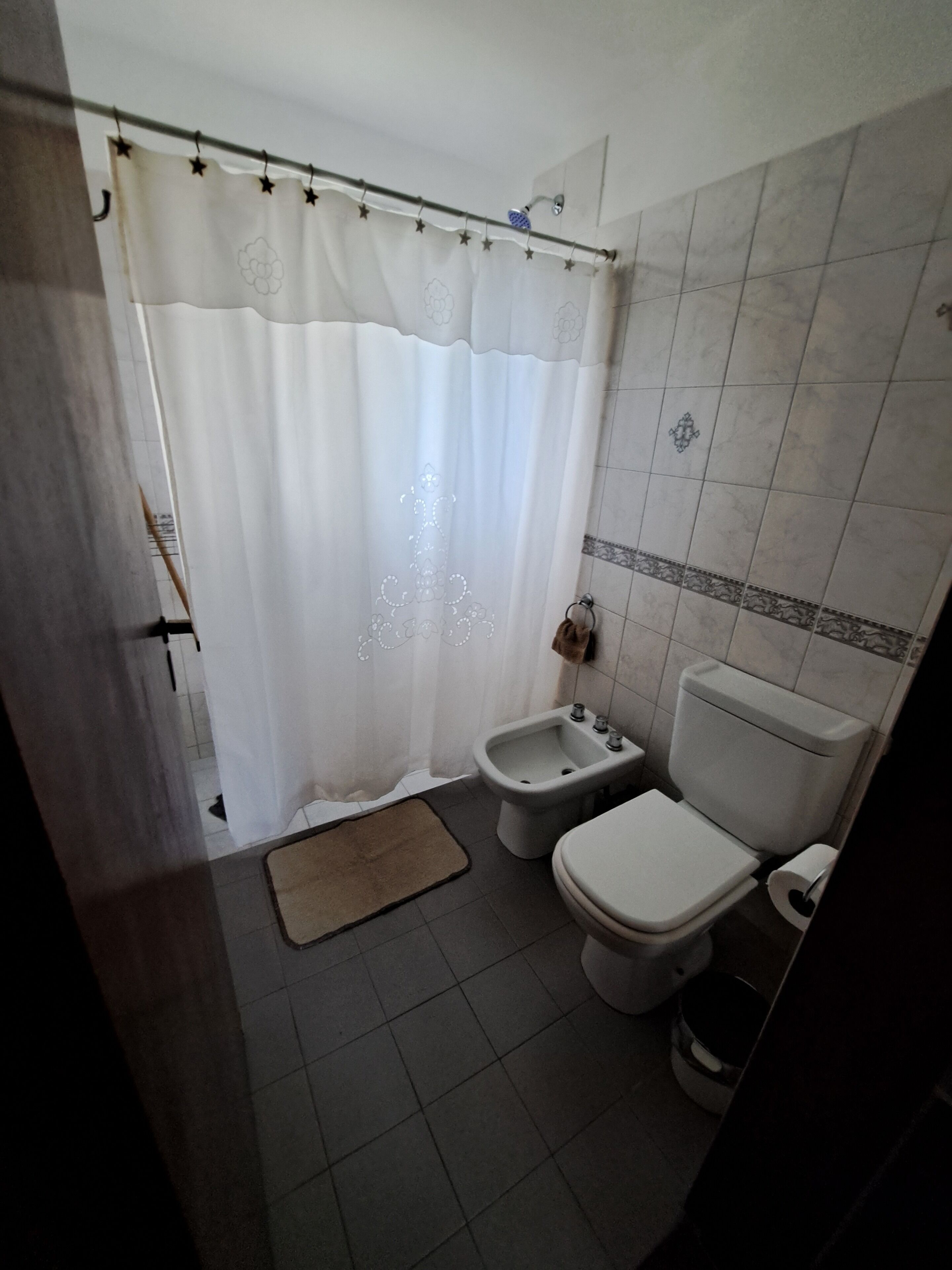 A bathroom with a shower curtain, a bidet, a toilet, and a small rug on the floor.