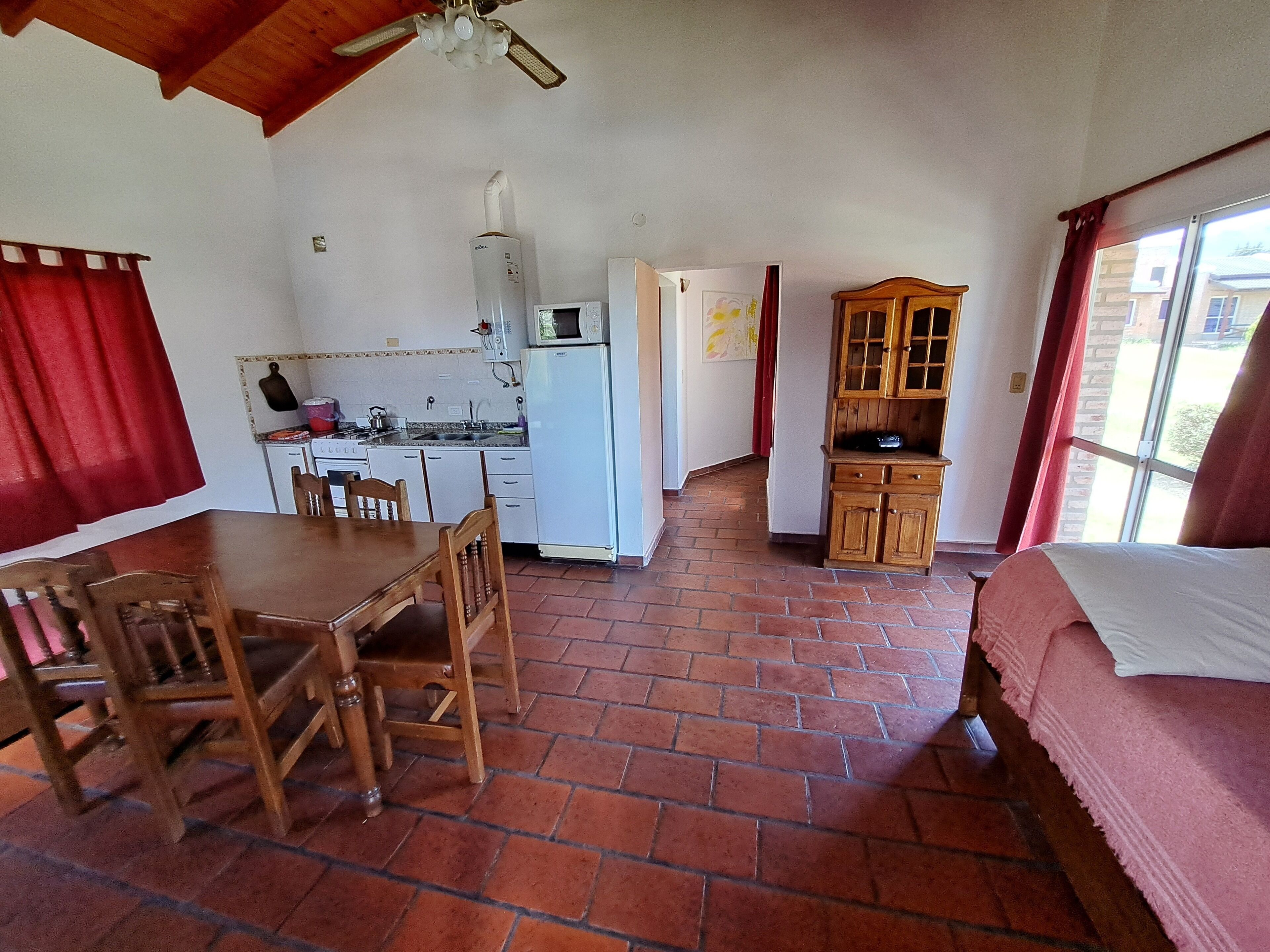 Habitació petita amb taula de menjador, cadires, llit, cuineta i un armari de fusta.