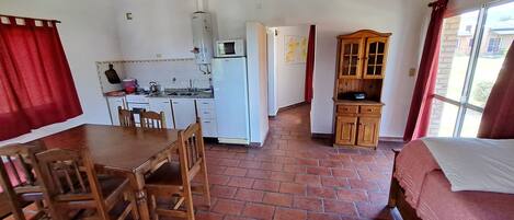 A compact room with a dining table, chairs, a bed, a kitchenette, and a wooden cabinet.