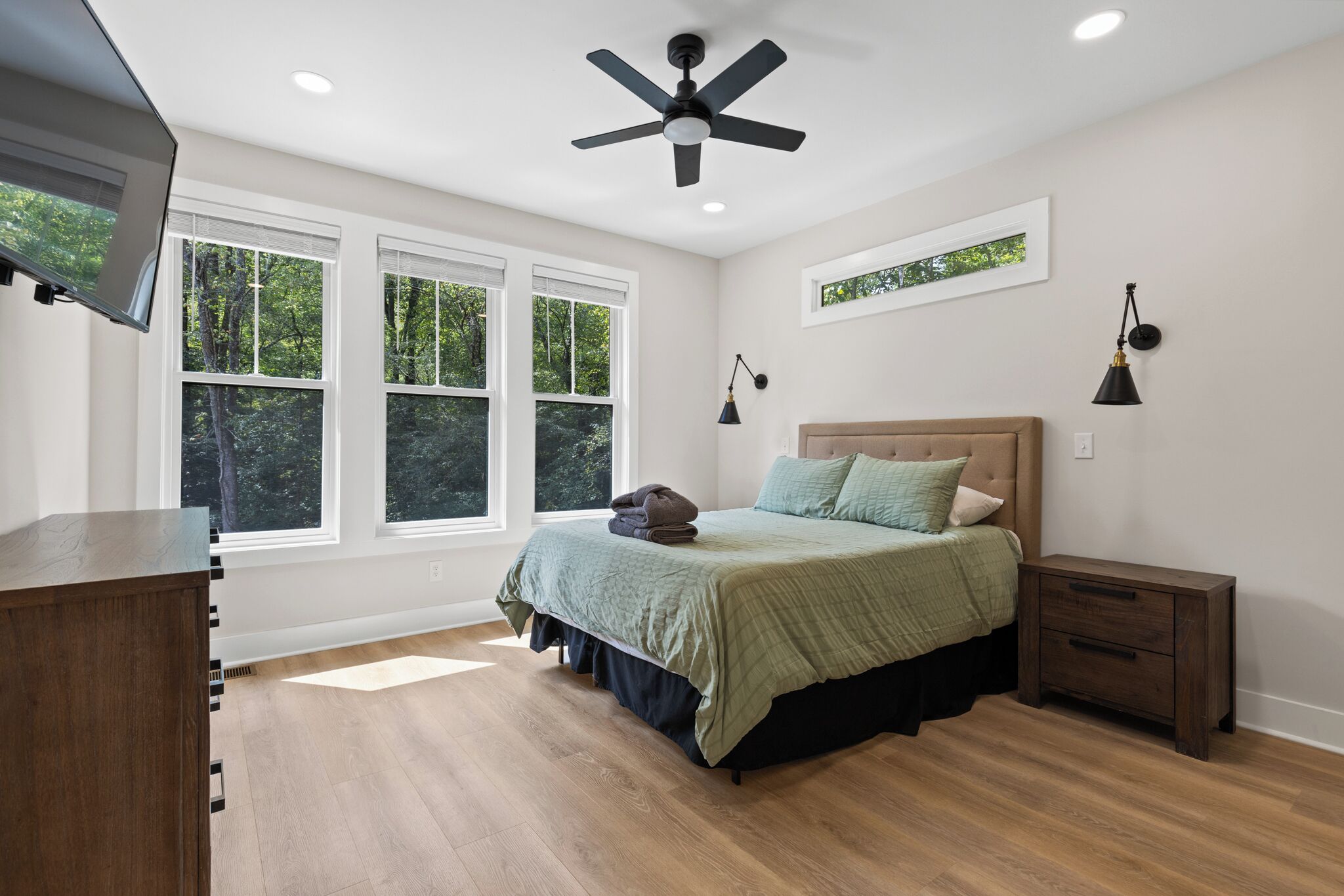 A bedroom with a bed, a ceiling fan, a TV, and a window with a view of trees.