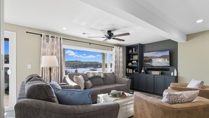 Una sala moderna con sofá, sillón y reposapiés, un ventilador de techo y televisor sobre una pared oscura.