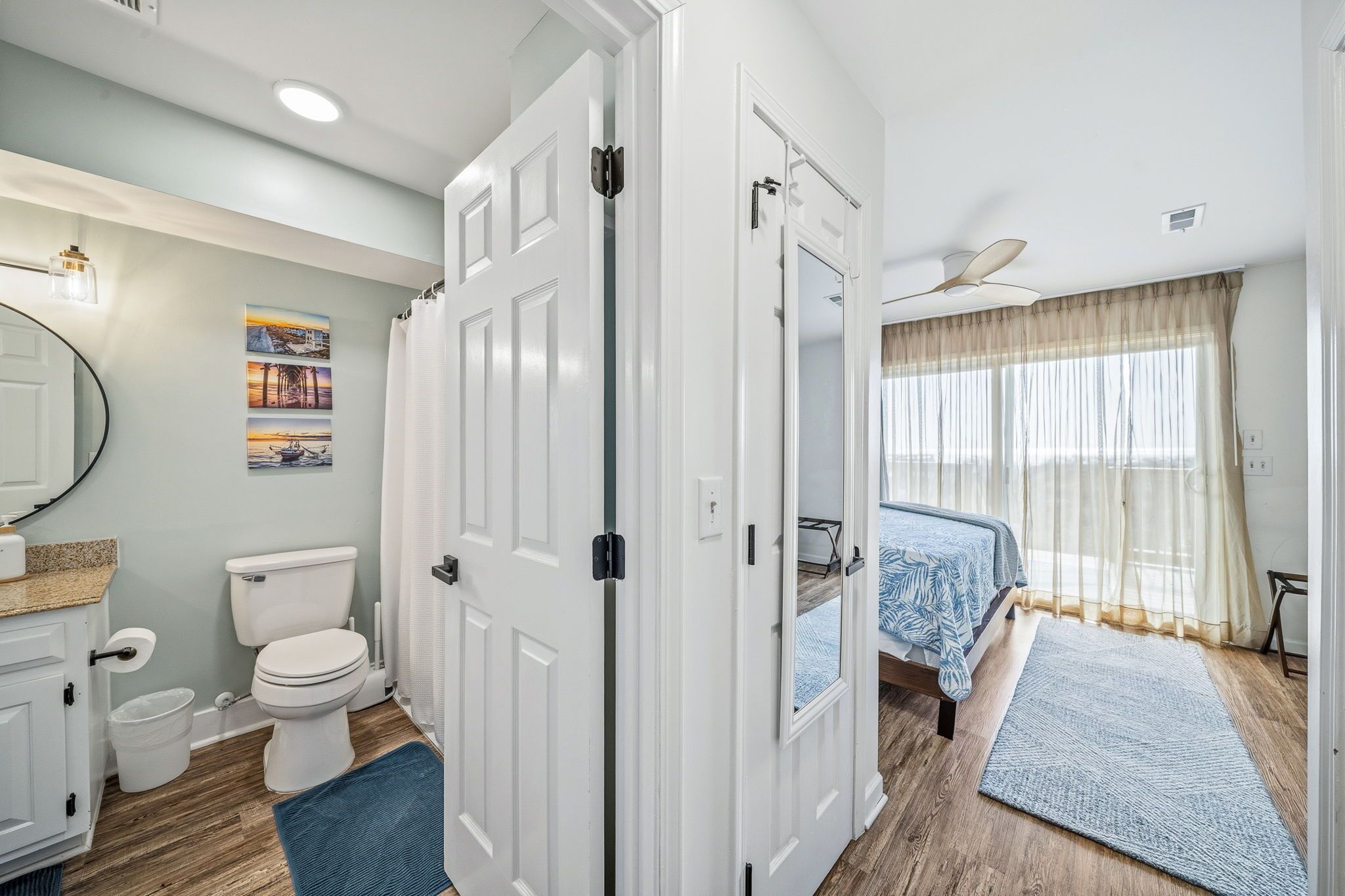 A bathroom with a toilet, sink, and mirror. There is a bed with a blue blanket and a window with curtains.